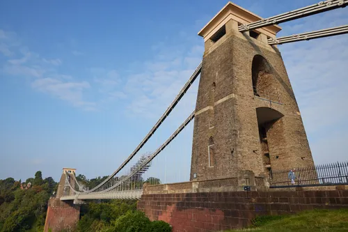 The image shows the Bristol Suspension Bridge in Bristol, England, United Kingdom, captured in the morning during late summer. This architecture photograph prominently features one of the stone towers and the suspension cables of the bridge, spanning across the Avon Gorge with lush green vegetation below. The Bristol Suspension Bridge is an iconic landmark recognized for its impressive engineering and design, and this view highlights its structural details as well as its integration with the surrounding landscape in Bristol.